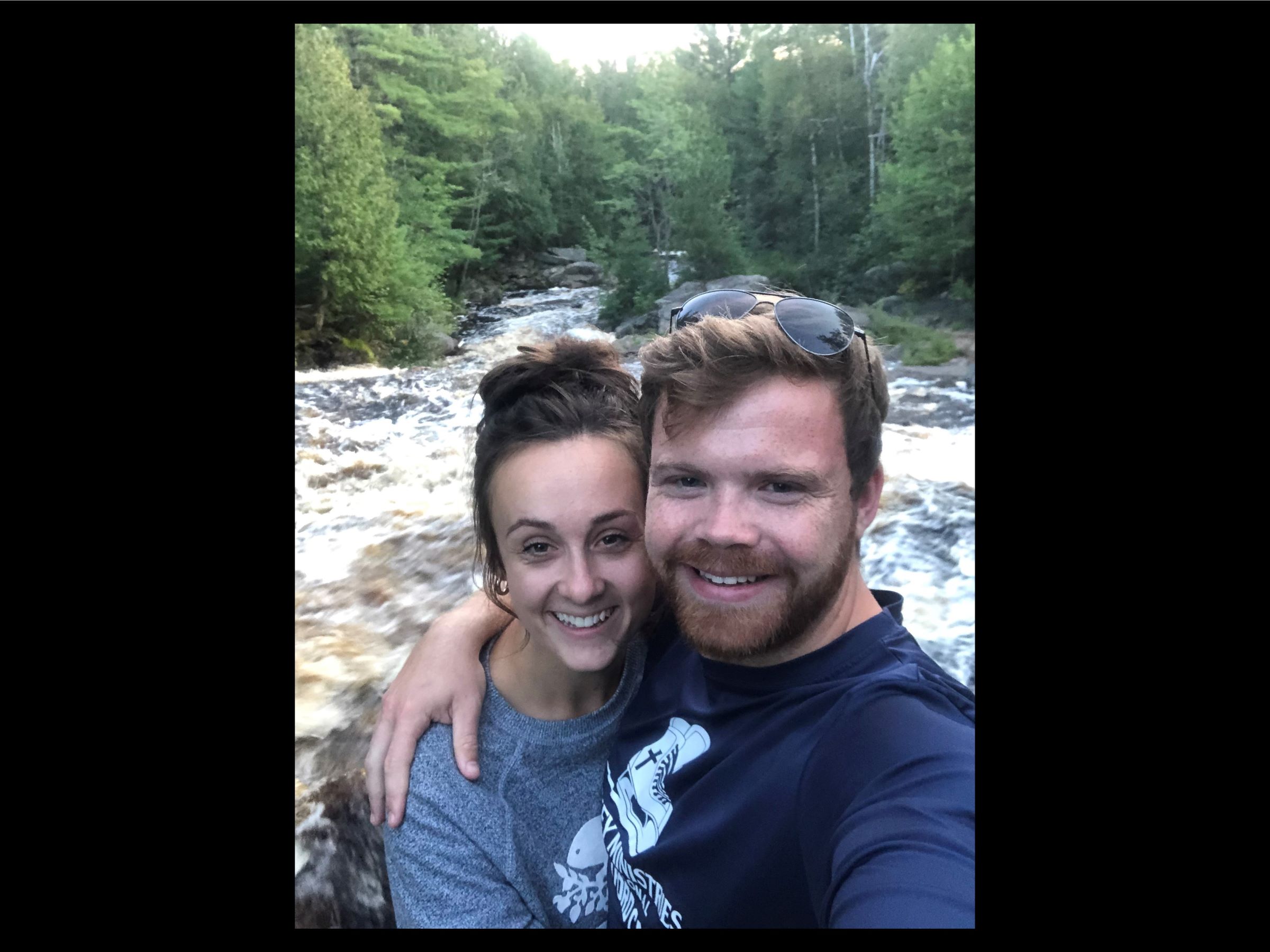 Homeowners Kara & Joshua are standing together with arms around each other. Behind them are lush trees and a flowing water stream.