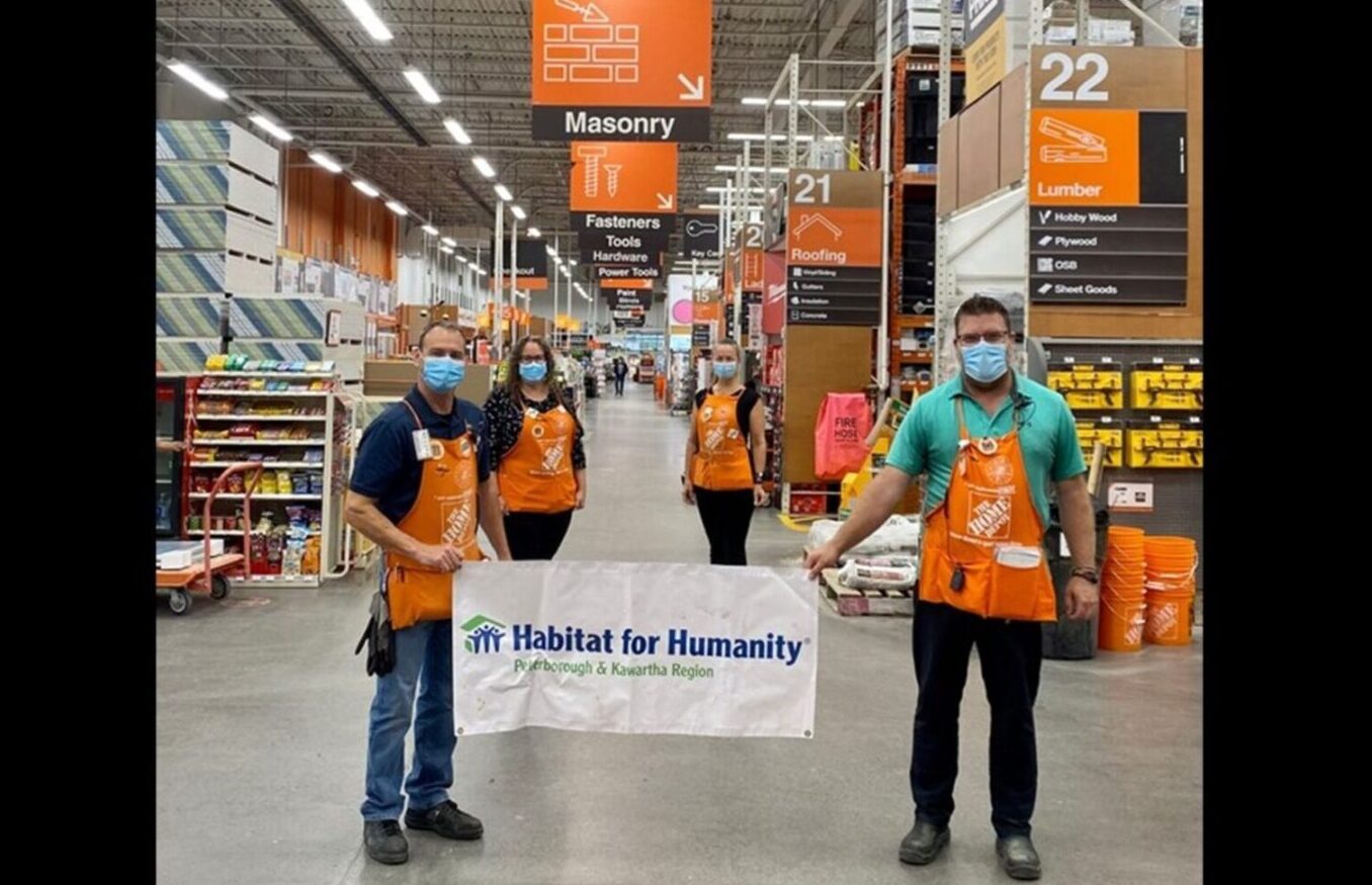 Four Home Depot employees standing inside the store holding a Habitat for Humanity banner. Behind them are Home Depot banners and shelves stocked with home building products.