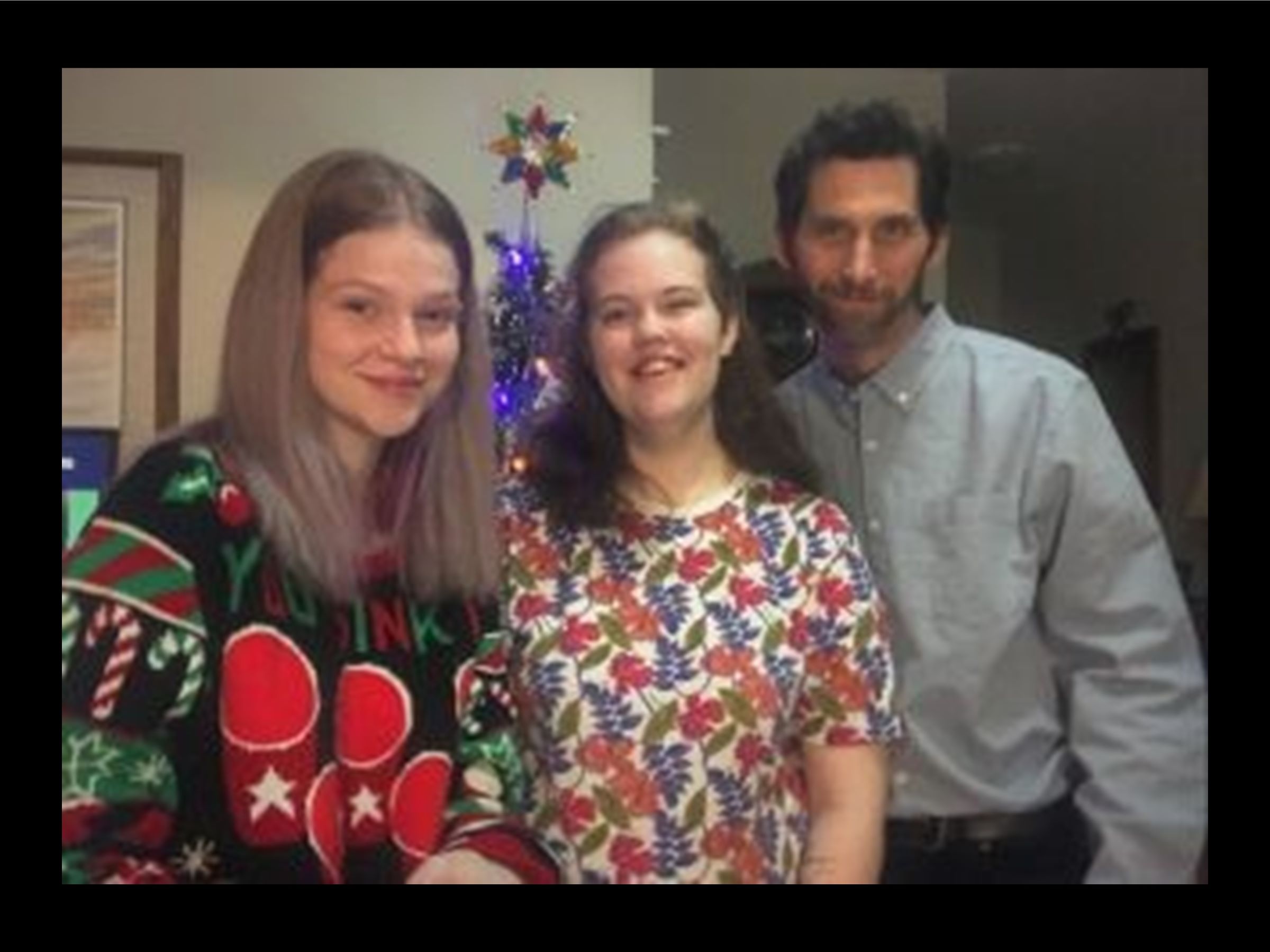 Two young women and father Deneil are all wearing christmas clothing and standing in front of a christmas tree, smiling.