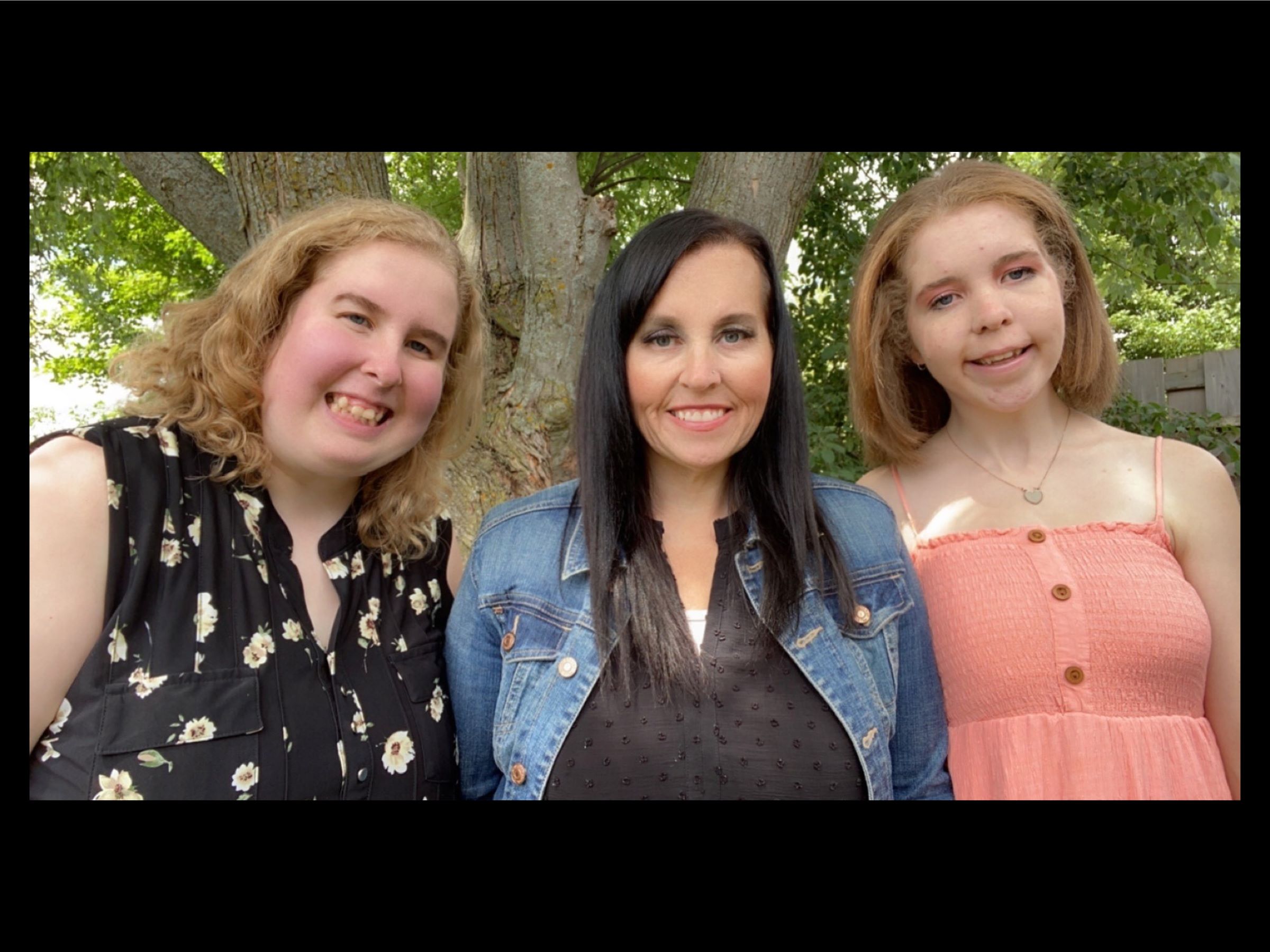 Mother Jody is standing in between her two daughters. The three women are standing outside in front of a lush green tree.