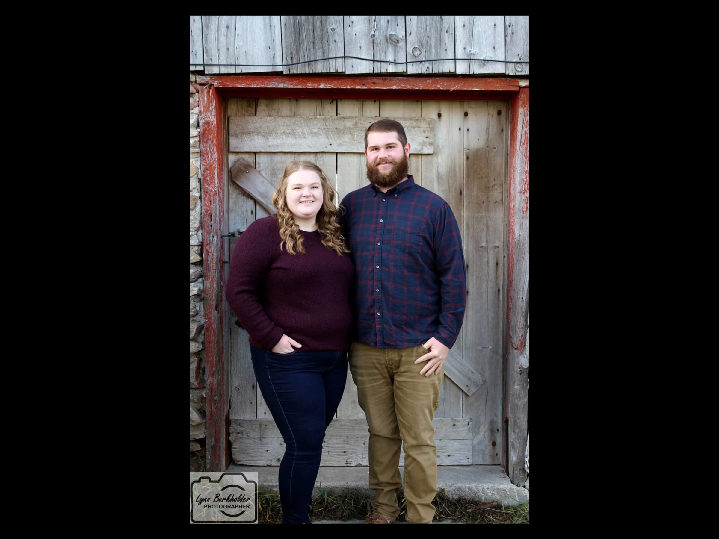 A young woman and man are standing outside in front of a barn door smiling, with one arm around each other and the other hand in their pocket.