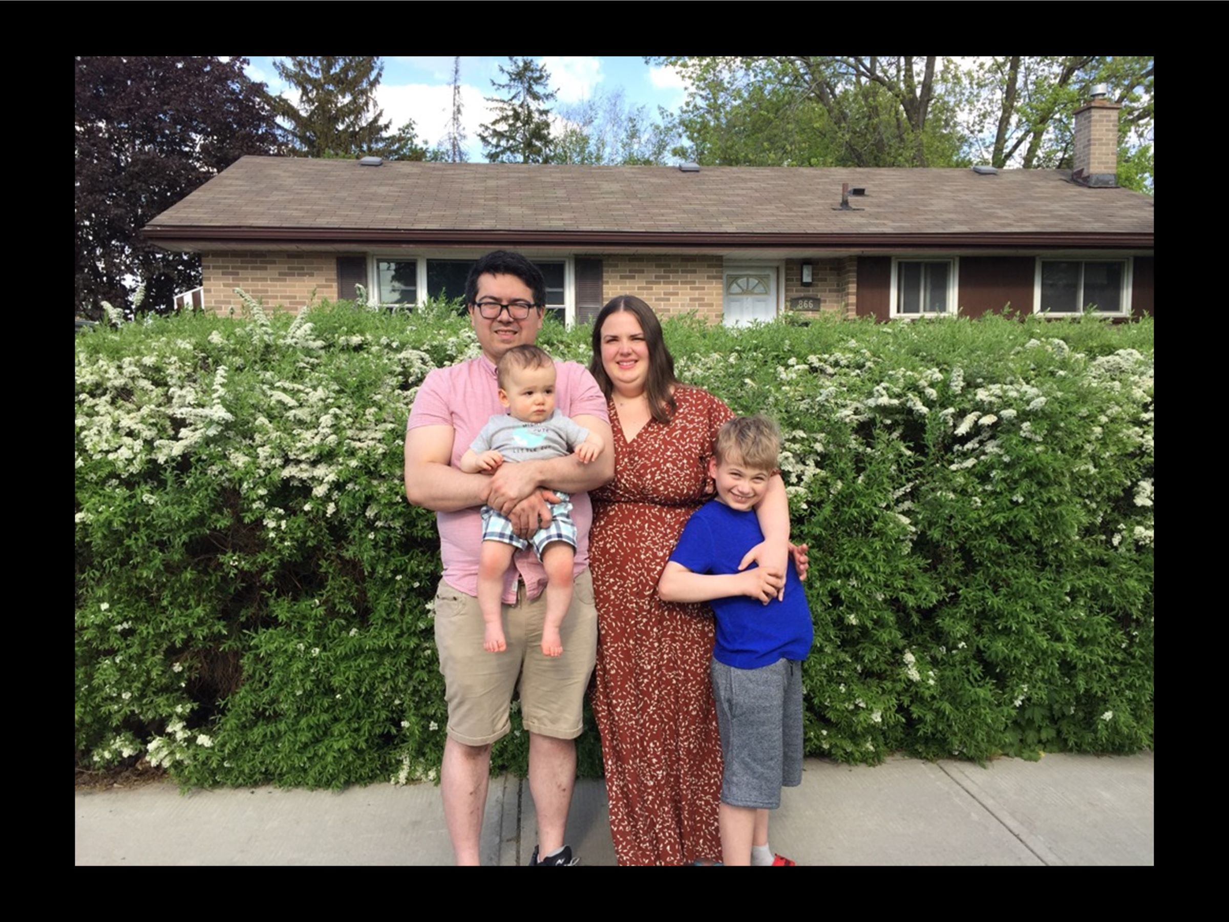 Parents Olivia & Paul standing together with their two young children. They are all standing in front of a large shrub in front of a brick house.