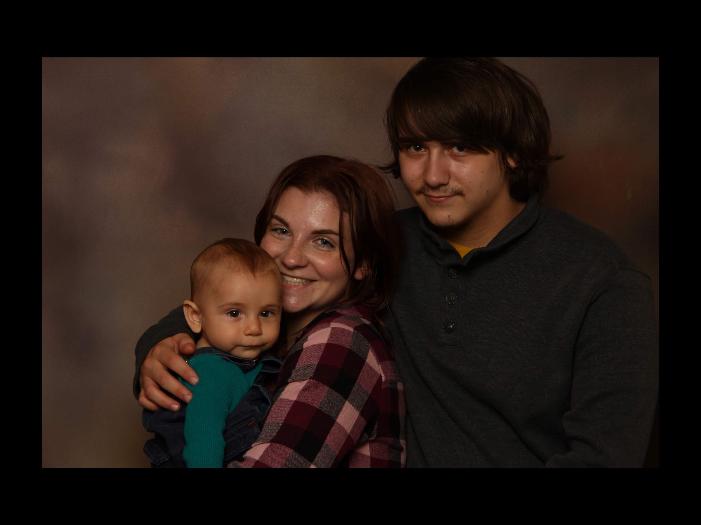 A young family, standing together smiling at the camera. Mother Sara has her arms around their young baby. Father Michael stands behind them.