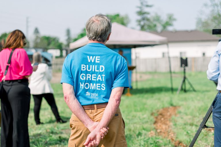 volunteer in a Habitat shirt that says "We build great homes" listening to the speakers at the Phase 2 Build Kick-Off