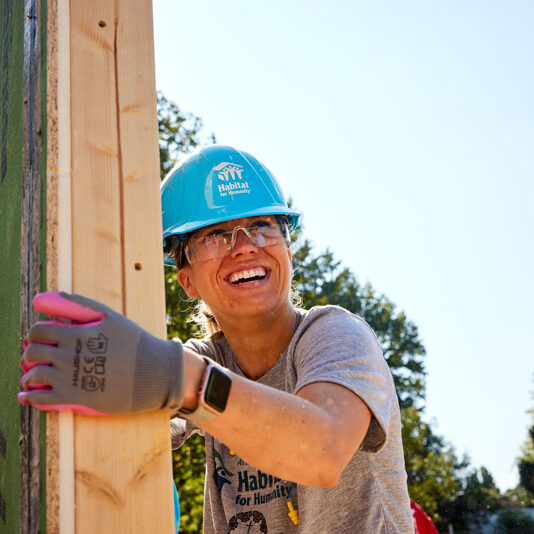 A woman in work gear, a blue Habitat construction hat, safety glasses and gloves, is carrying a piece of lumber with a bright, joyful smile on her face